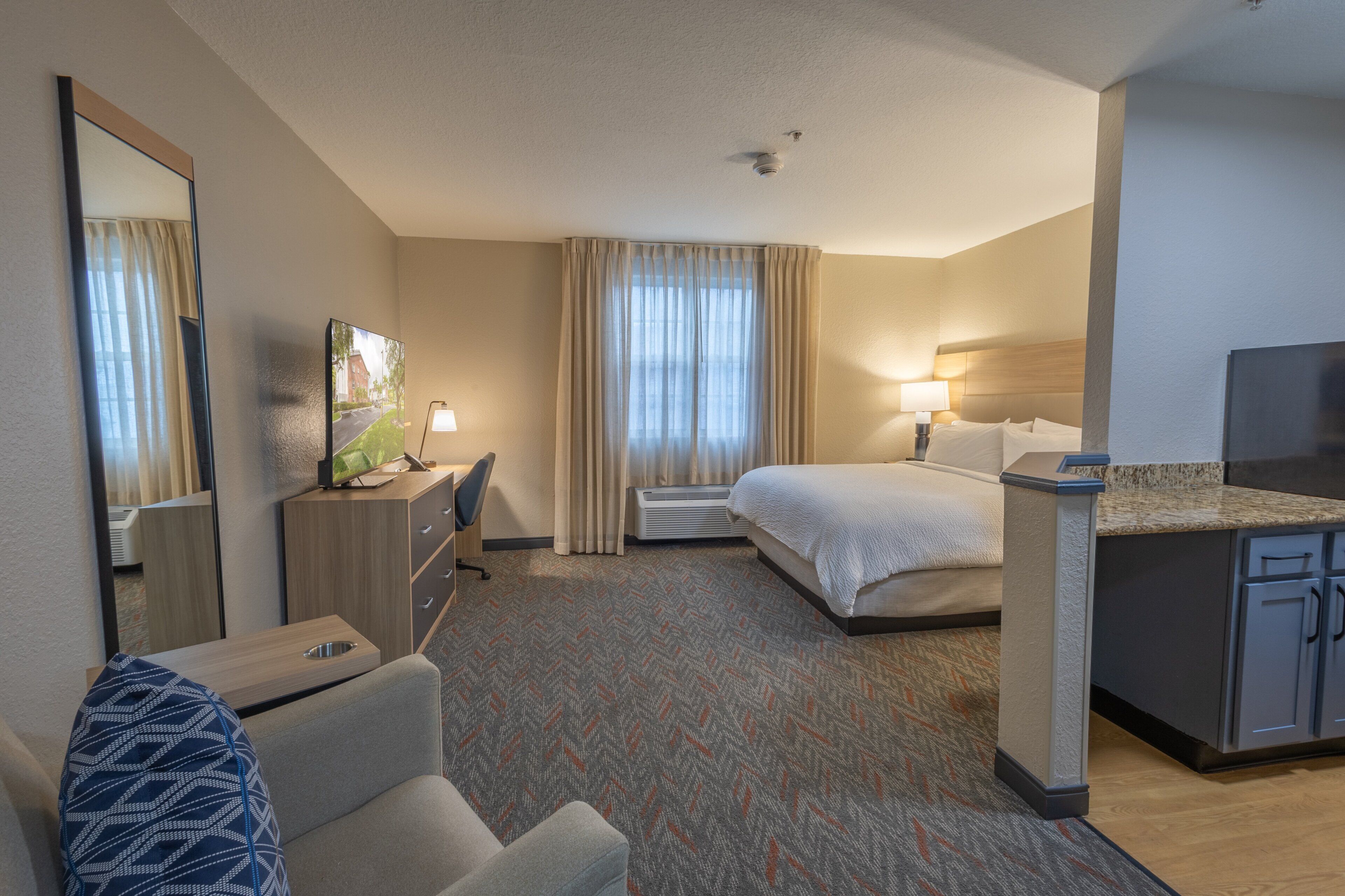 A hotel room with a bed, a desk with a chair, a television, a mirror, and a kitchenette.