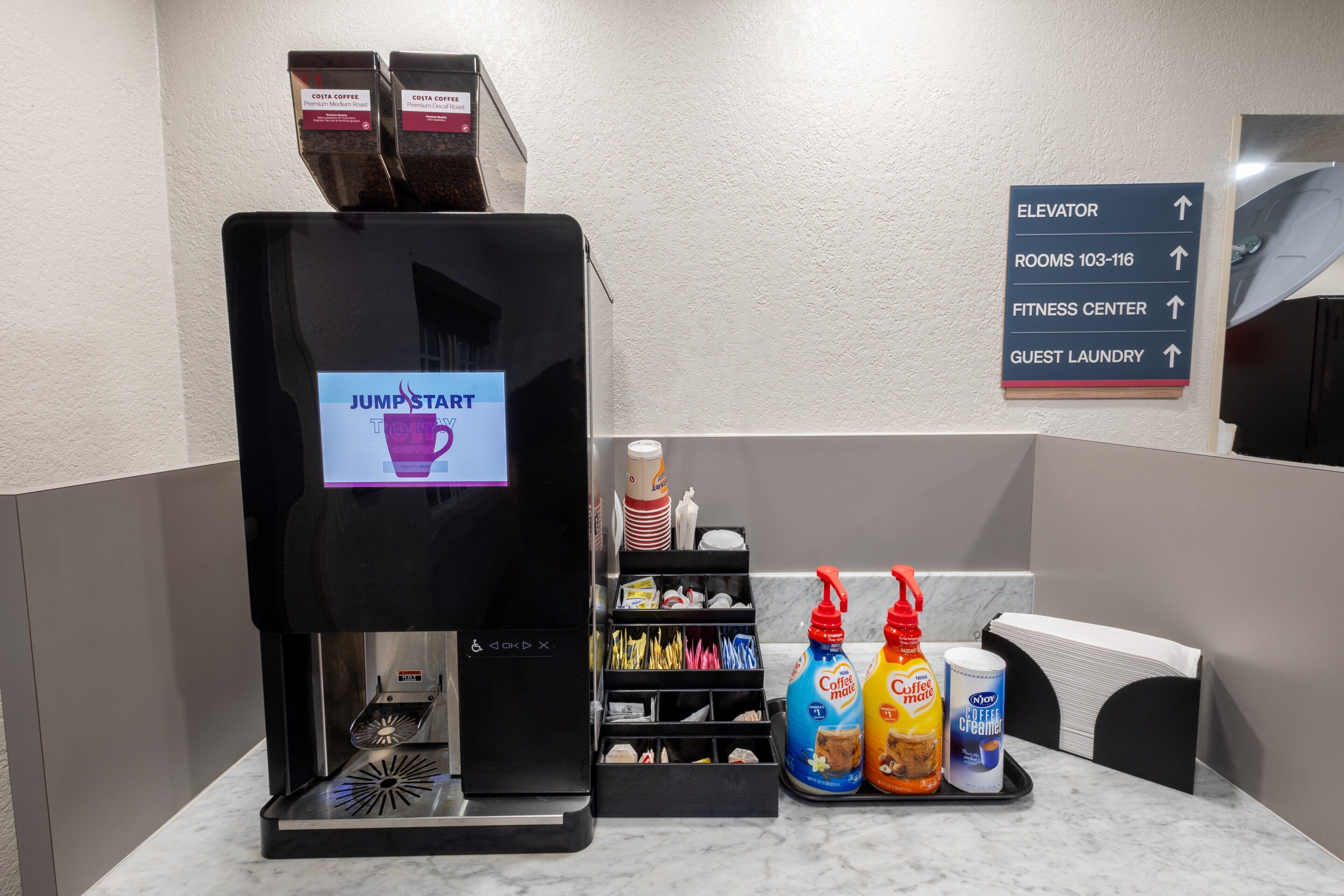 A coffee machine with a digital display, a sign indicating directions to the elevator, rooms, fitness center, and guest laundry, and a tray with cleaning supplies.