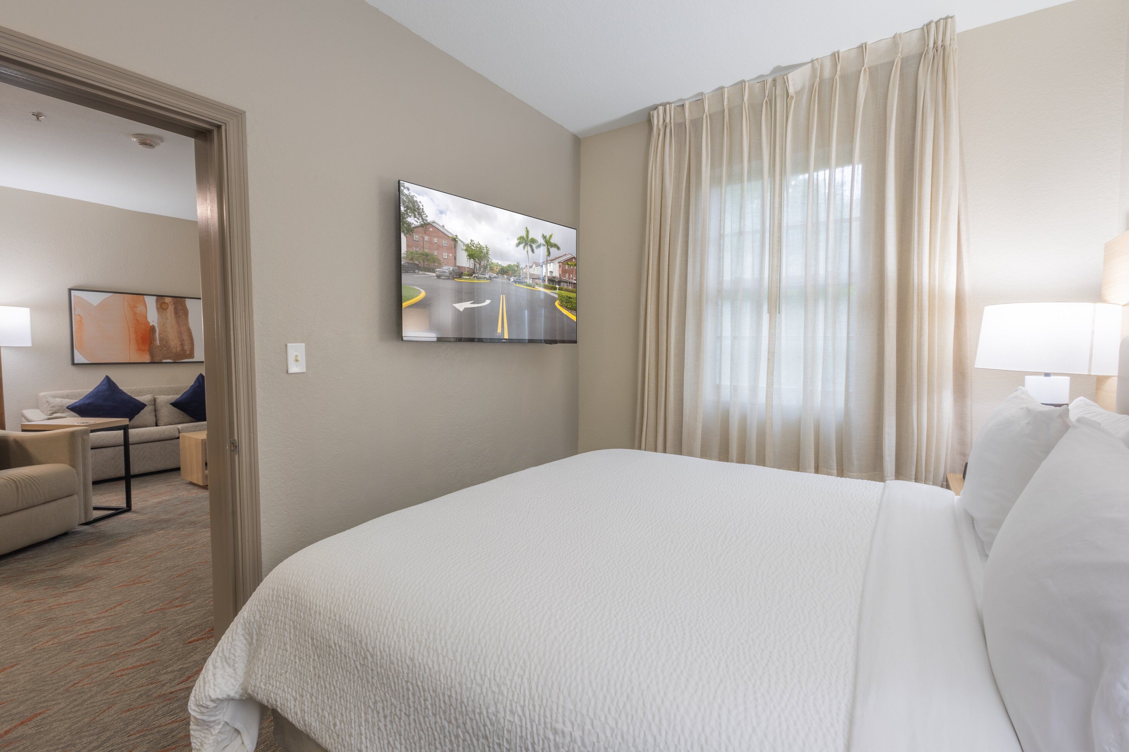 A hotel room with a bed, a TV mounted on the wall, a window with curtains, and a sofa with pillows.