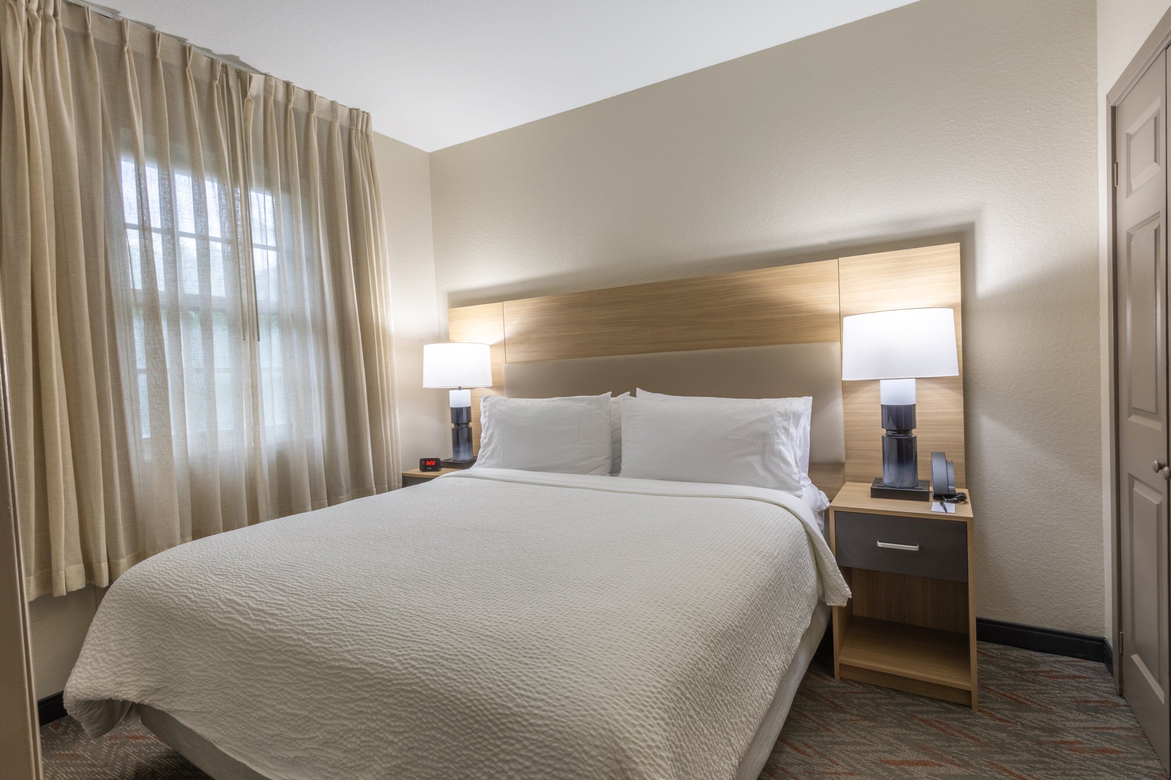 Een hotelkamer met een groot bed, twee nachtkastjes met lampen, een bijzettafeltje en een raam met gordijnen.