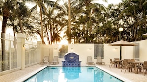 A swimming pool with a blue tiled area, surrounded by palm trees and a white fence.