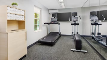 Een fitnessruimte met loopband, elliptische trainer en hometrainer, een houten kast met handdoeken, en een raam met uitzicht op groen.