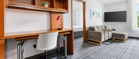 A modern hotel room with a wooden desk, a white office chair, a gray sofa, and a small table.