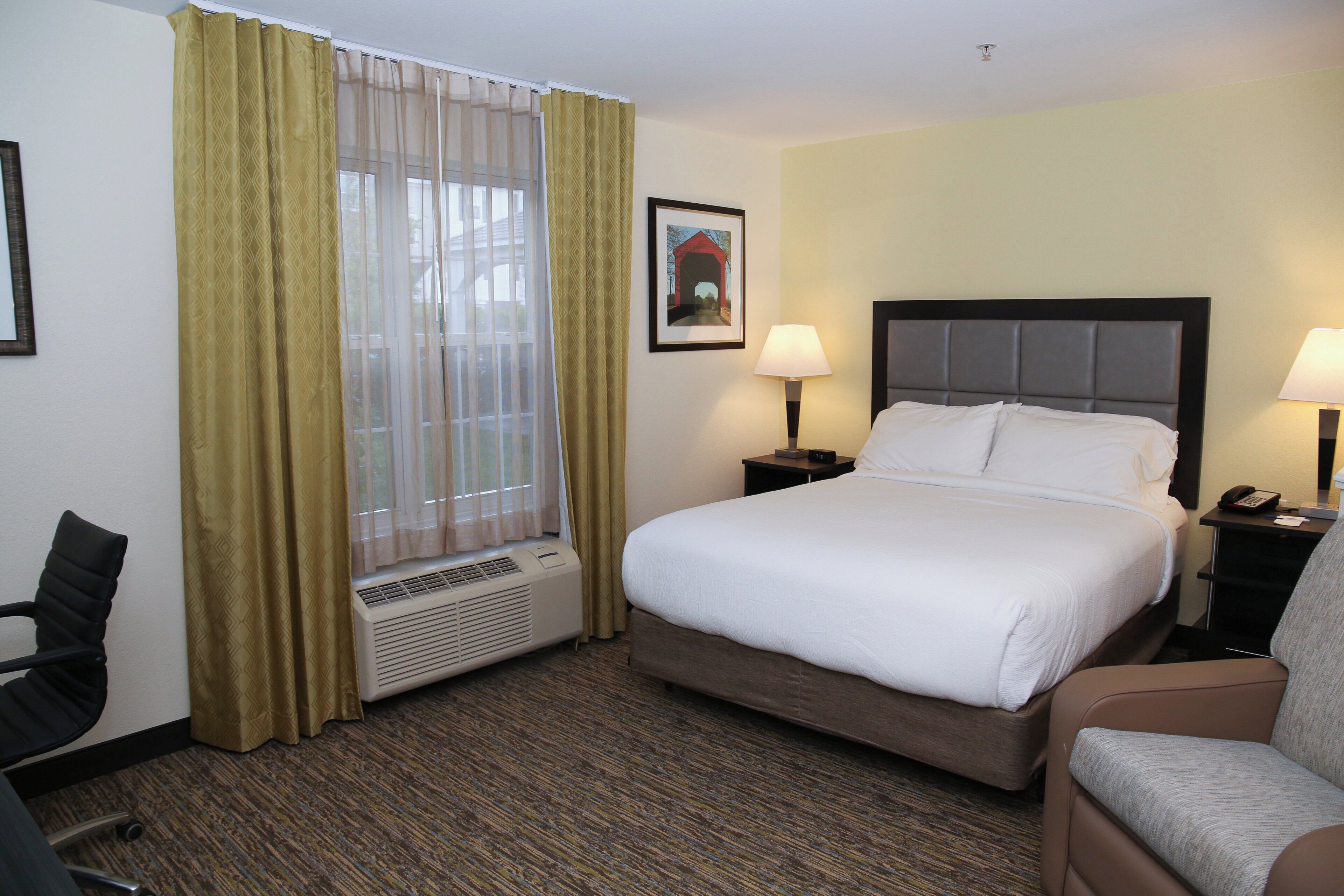 Habitación de hotel con cama, dos lámparas de noche, ventana con cortinas, calefactor, una silla e imagen enmarcada en la pared.