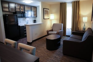 Habitación de hotel funcional con kitchenette, zona de estar con sofá y sillones, una mesita redonda y una lámpara.