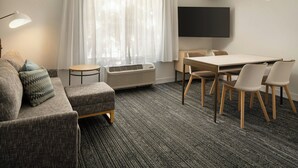 A modern living room with a grey sofa, a small round table, a flat-screen TV, and a dining area with chairs.