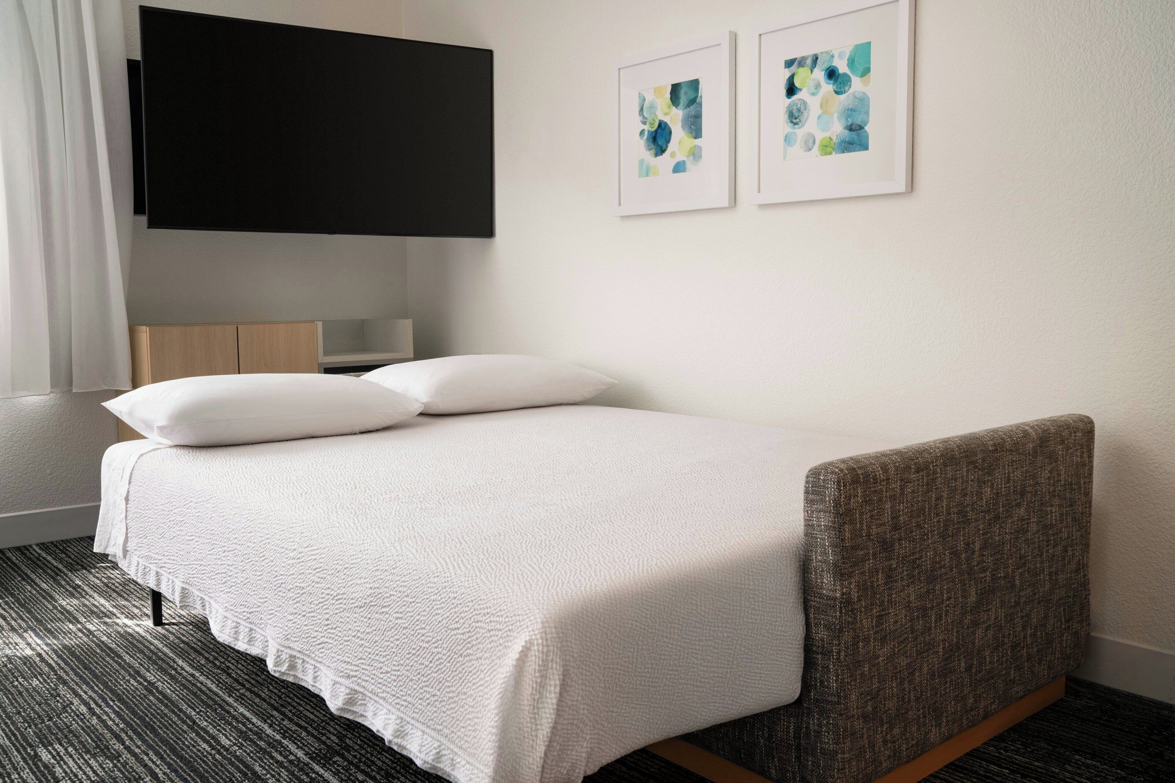 A neatly made bed with a white bedspread and pillows, a flat-screen TV mounted on the wall, and two framed abstract art pieces on the wall.