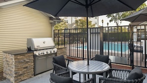 An outdoor patio with a grill, wicker chairs, and a pool in the background.