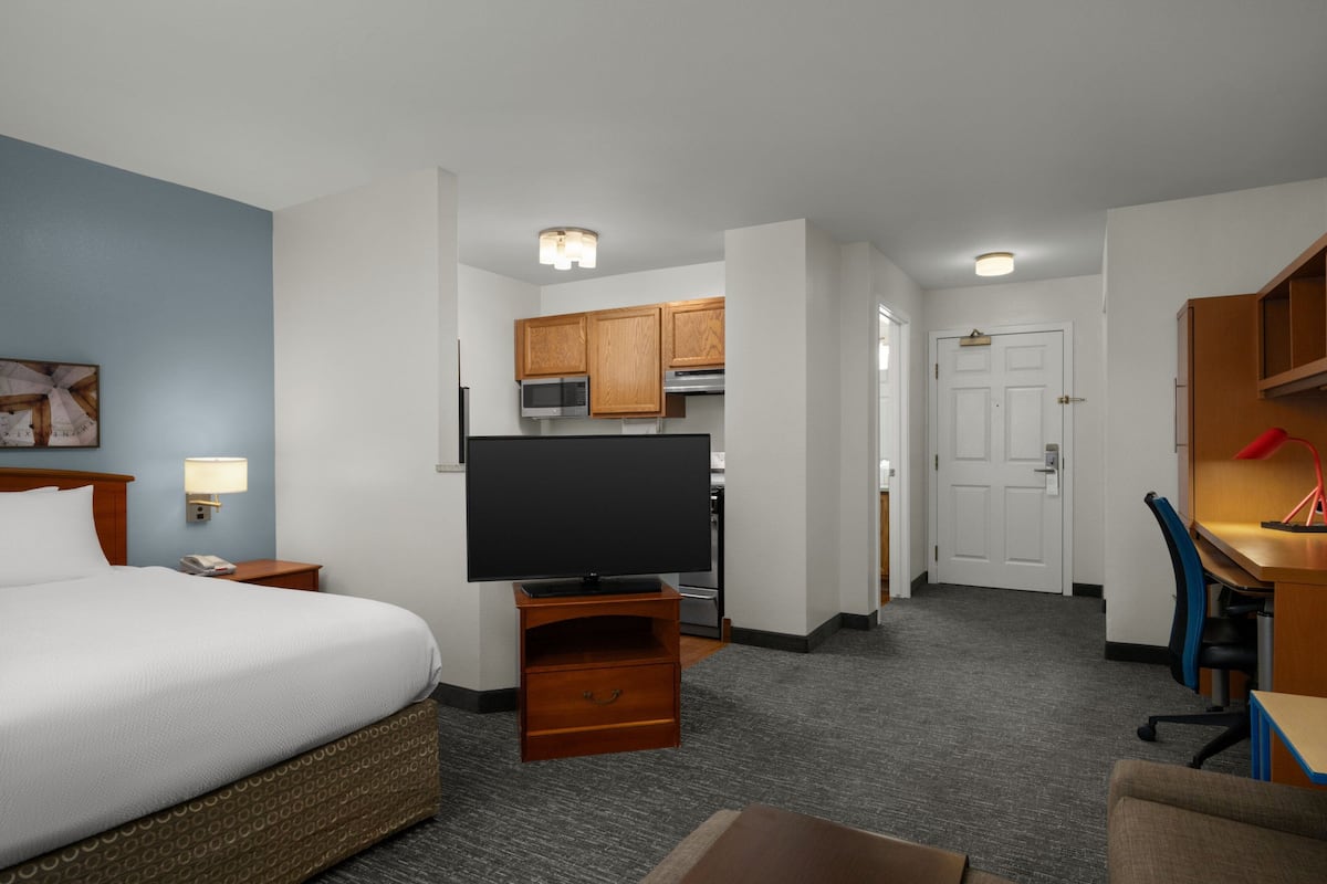 Habitación de hotel con cama, televisor sobre un mueble de madera, escritorio con silla, kitchenette y una puerta que da a otra habitación.