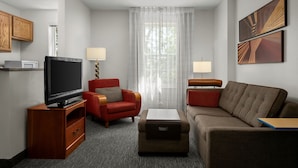 A modern hotel room with a flat-screen TV, a red armchair, a brown sofa, and a large window with sheer curtains.
