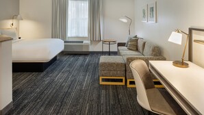 A hotel room with a bed, a sofa, a desk, and a chair. There is a window with curtains, a lamp, and an air conditioning unit. - Towneplace Suites By Marriott Findlay (Findlay)