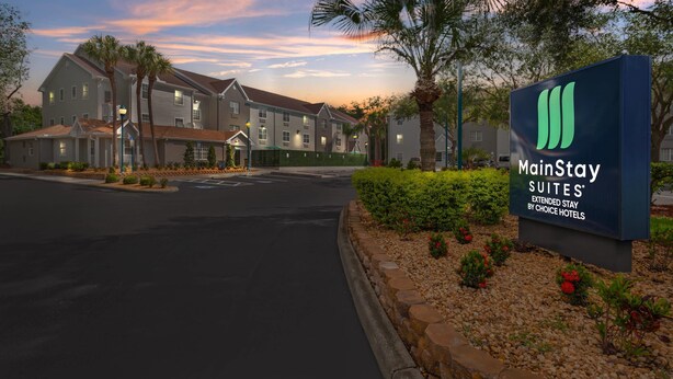 A MainStay Suites hotel entrance with a sign, palm trees, and a road leading to the building. - MainStay Suites Tampa Moffitt-USF (Tampa)