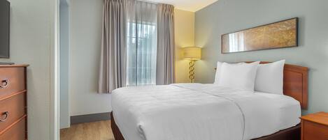 A hotel room with a large bed, a wooden nightstand, a desk with a lamp, a TV, and a window with curtains.