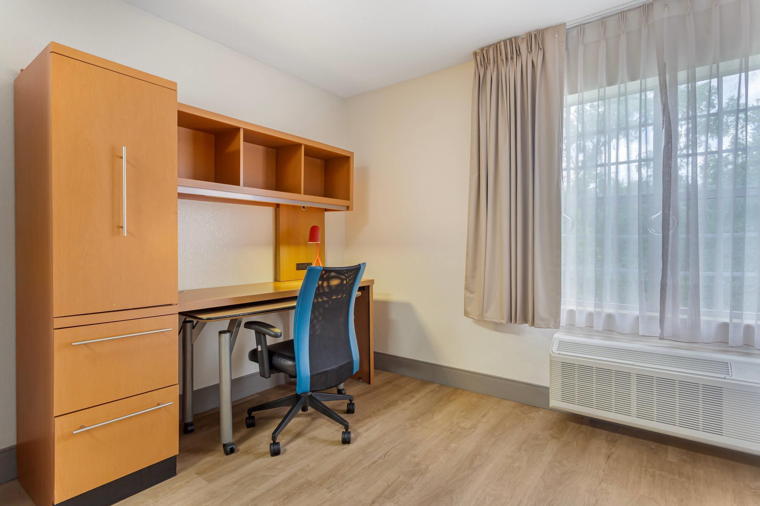 Una habitación con un escritorio de madera, una silla, un armario y una ventana con cortinas.