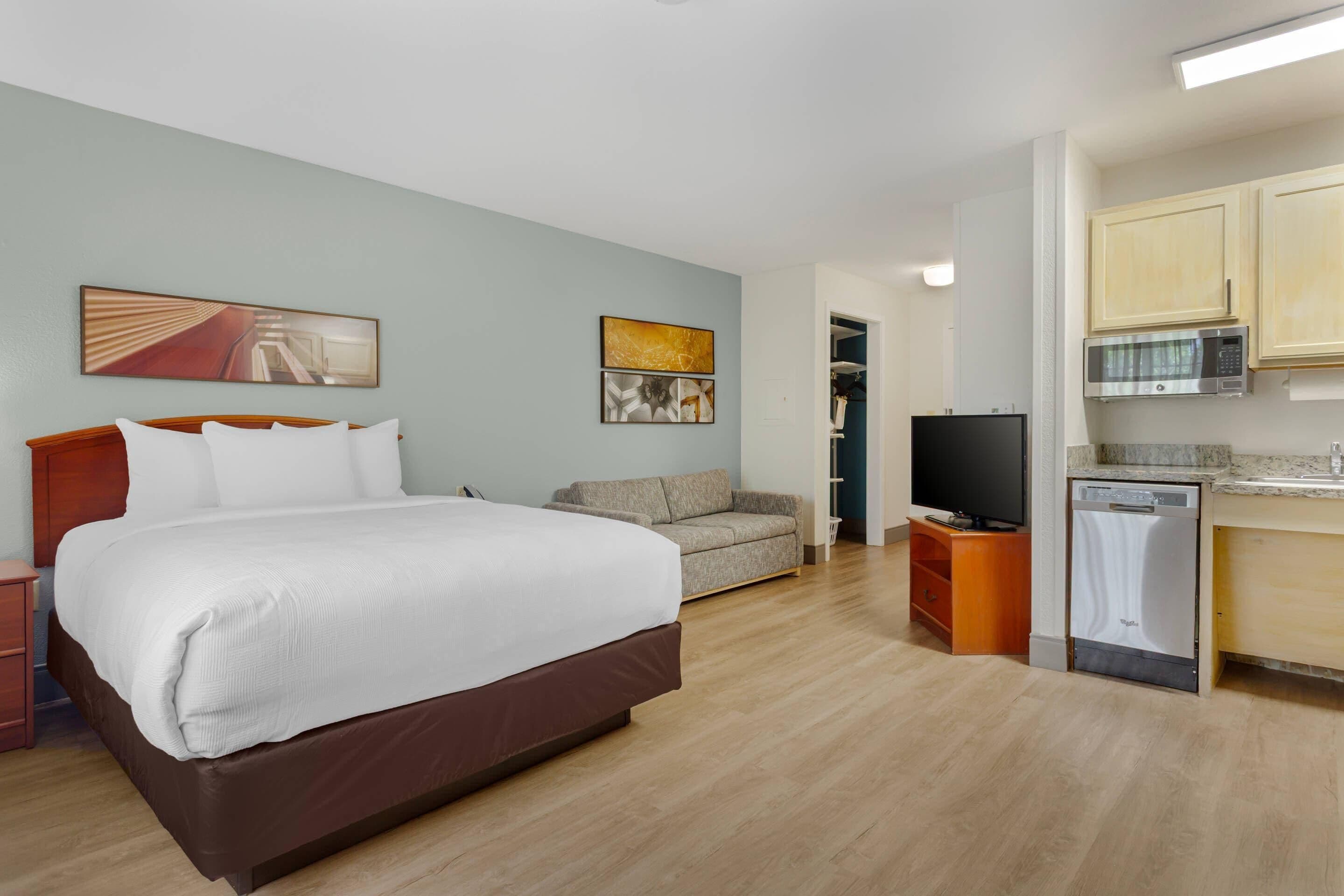 A hotel room with a large bed, a sofa, a television, and a kitchen area with a microwave and dishwasher.