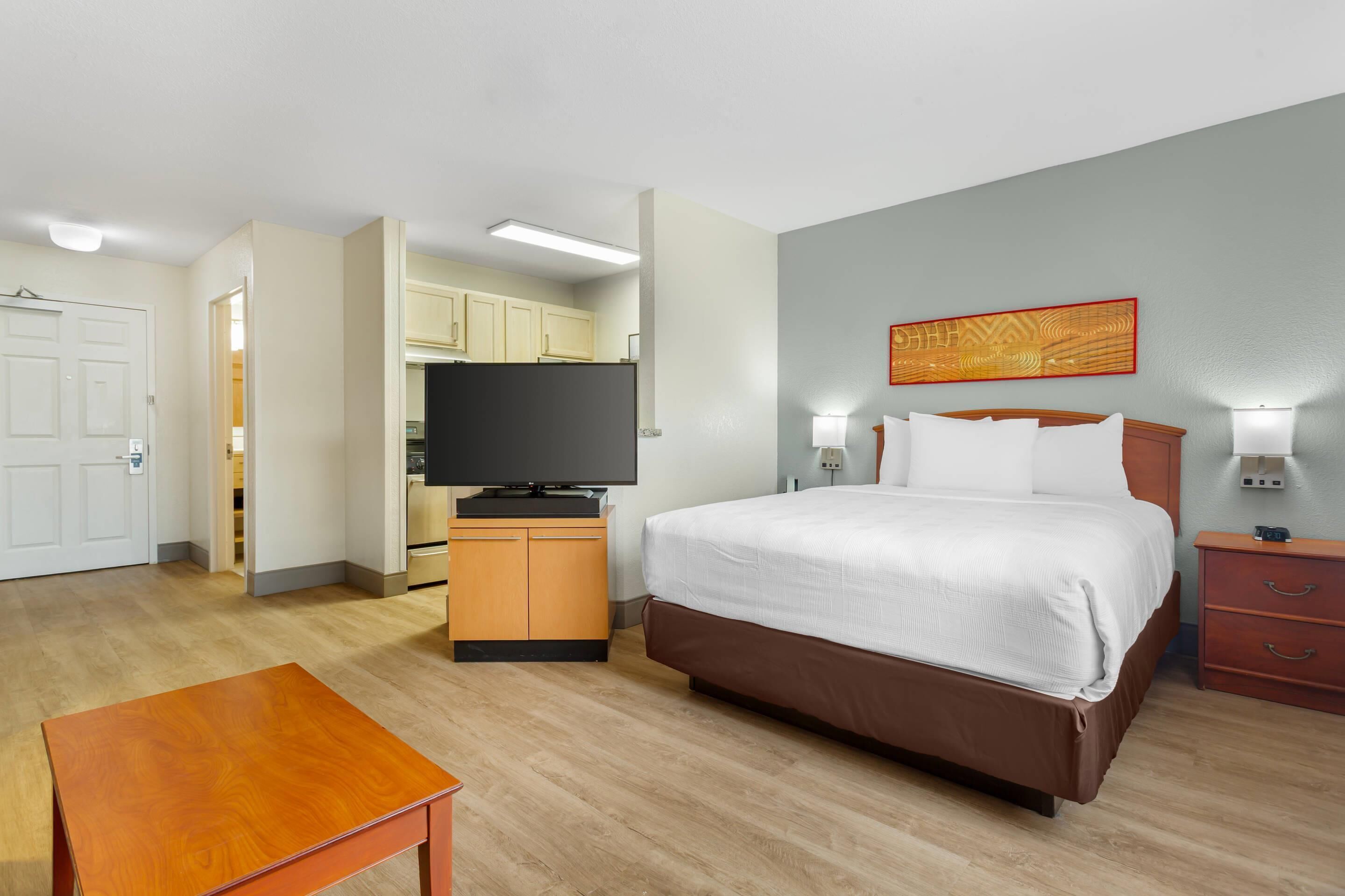Una habitación de hotel con cama, lámparas de noche, televisor, mesa de madera y kitchenette.