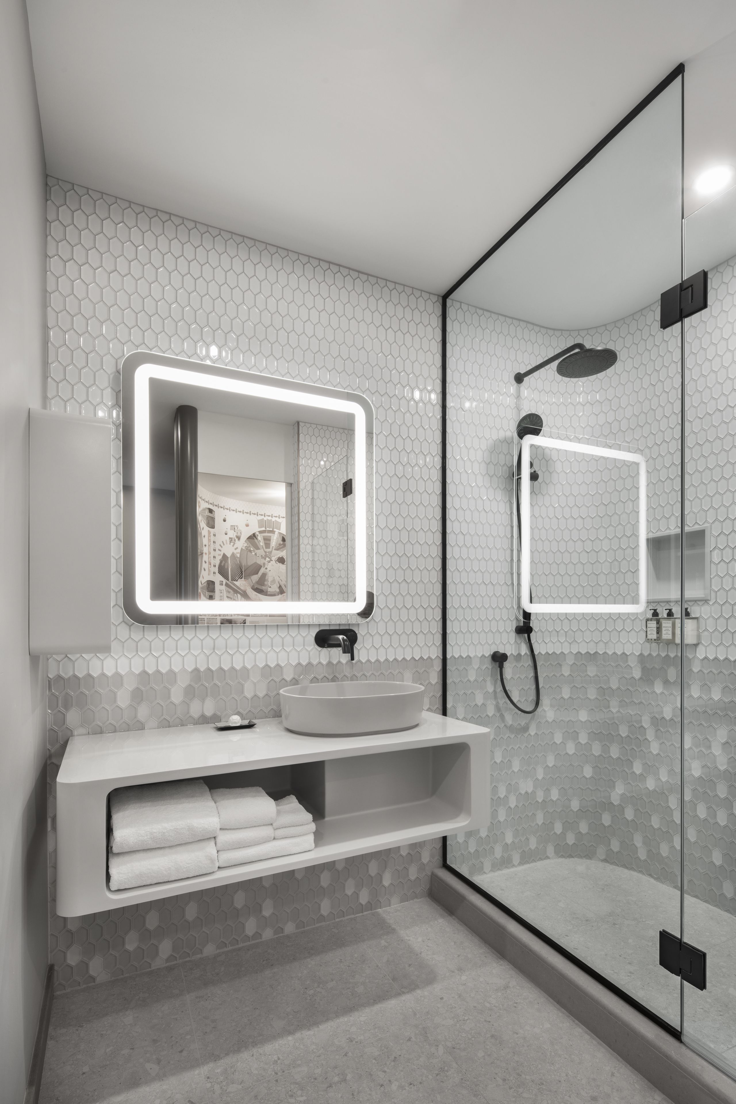 A modern bathroom with a glass shower enclosure, a large mirror with LED lighting, and a white vanity with a sink and towel storage.