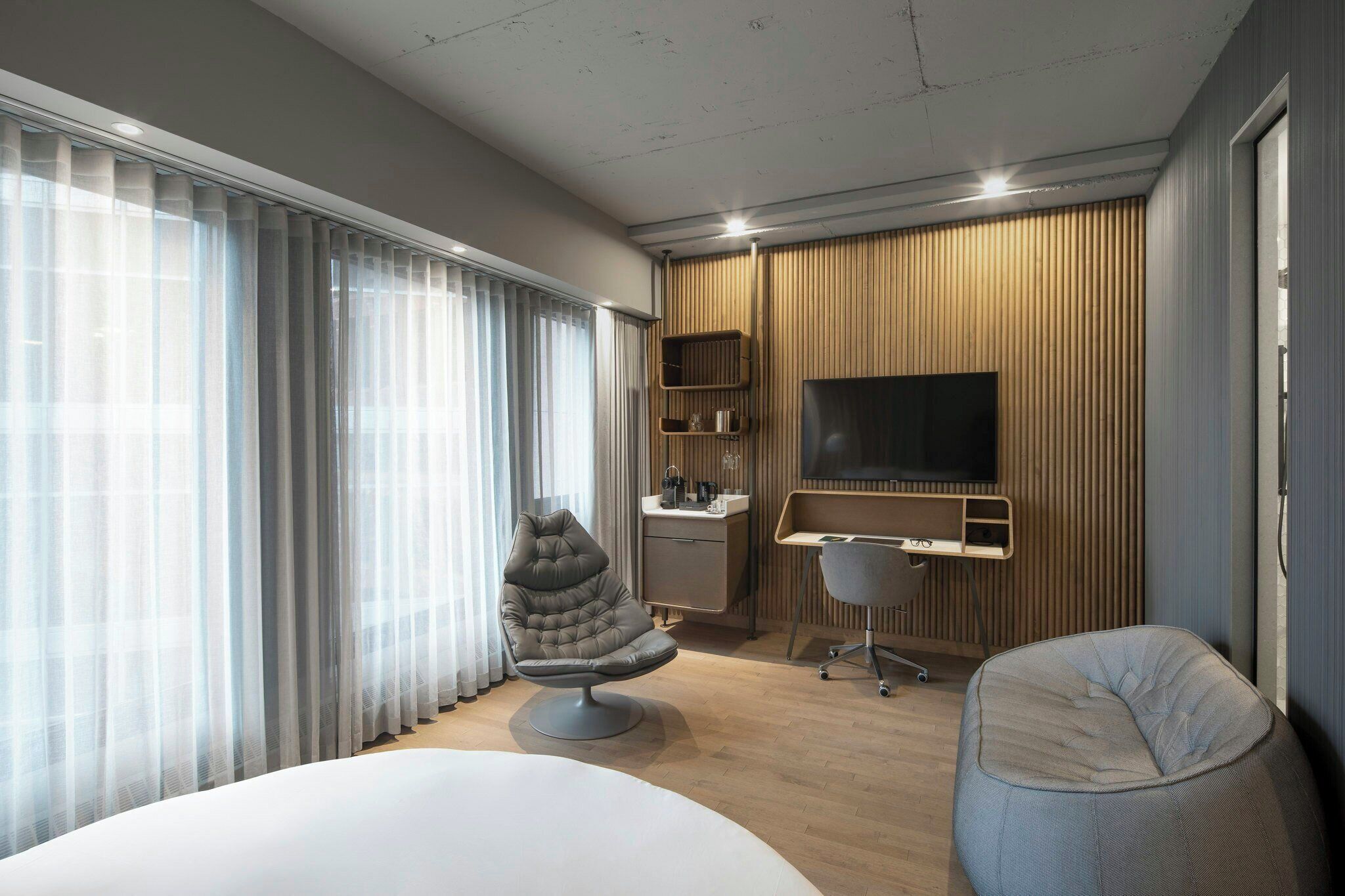 A modern hotel room with a large bed, a comfortable armchair, a desk with a chair, a flat-screen TV mounted on the wall, and a large window with sheer curtains.