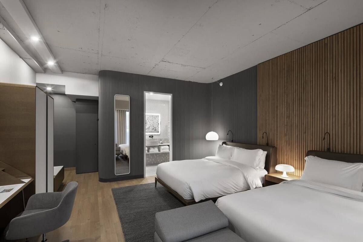 A modern hotel room with two beds, a desk, and a chair. There is a bathroom visible through an open door.