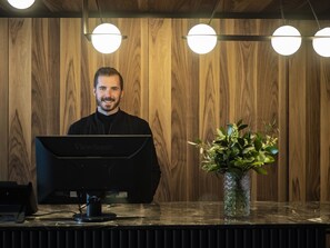 Un mostrador de recepción con un monitor de computadora, un florero con plantas verdes y paneles de madera en el fondo.