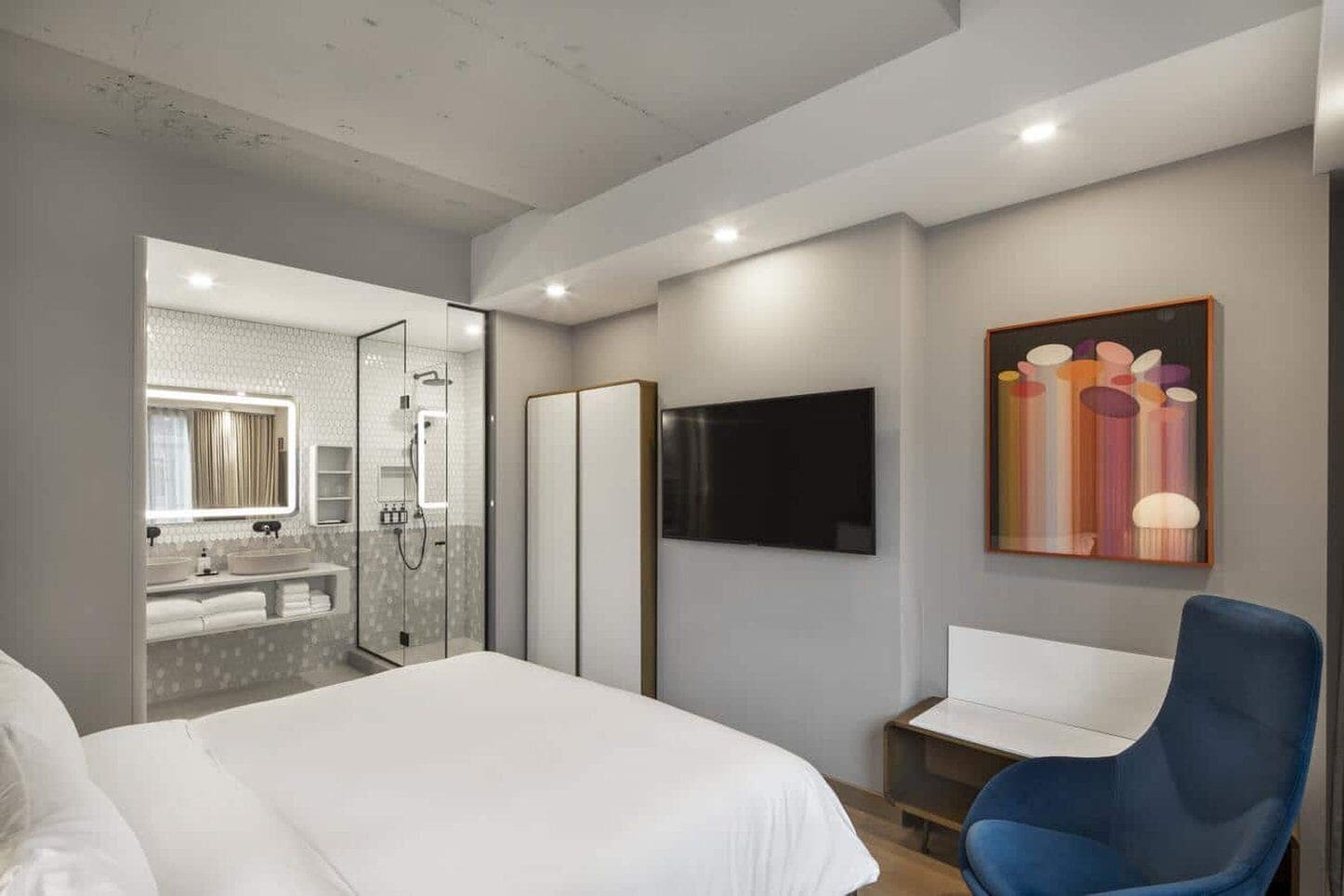 Una habitación de hotel moderna con una cama grande, zona de ducha, televisor y una silla azul.