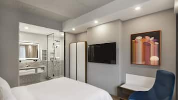 Una habitación de hotel moderna con una cama grande, zona de ducha, televisor y una silla azul.