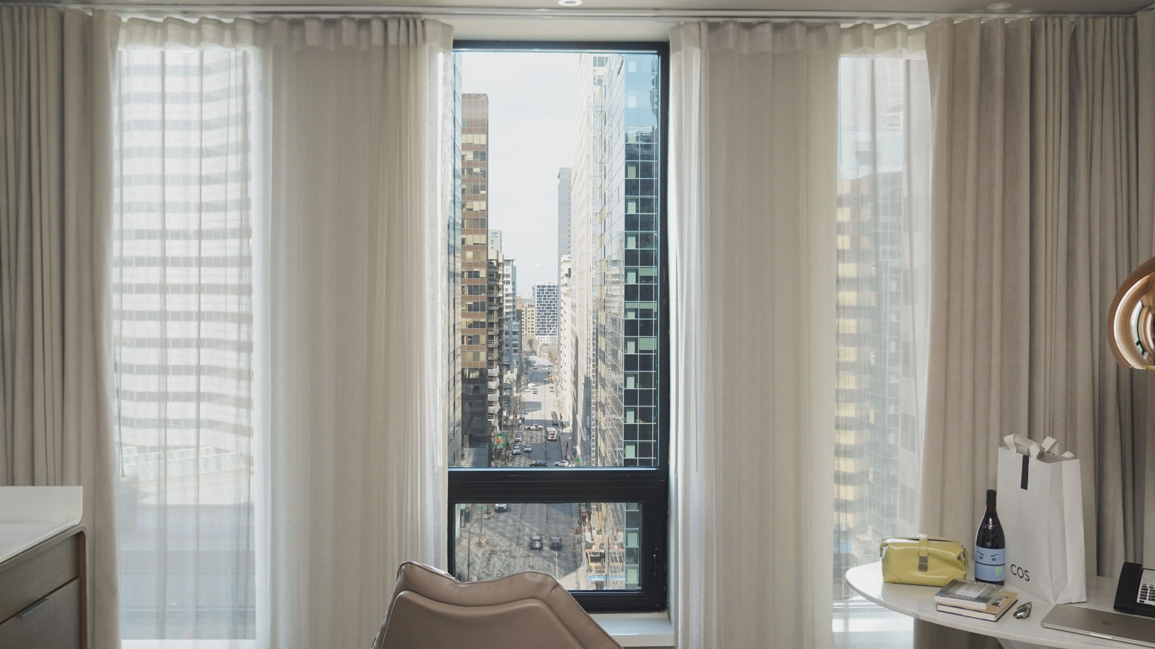 Une chambre d’hôtel avec une grande fenêtre, un bureau avec un ordinateur portable, une bouteille et un sac.