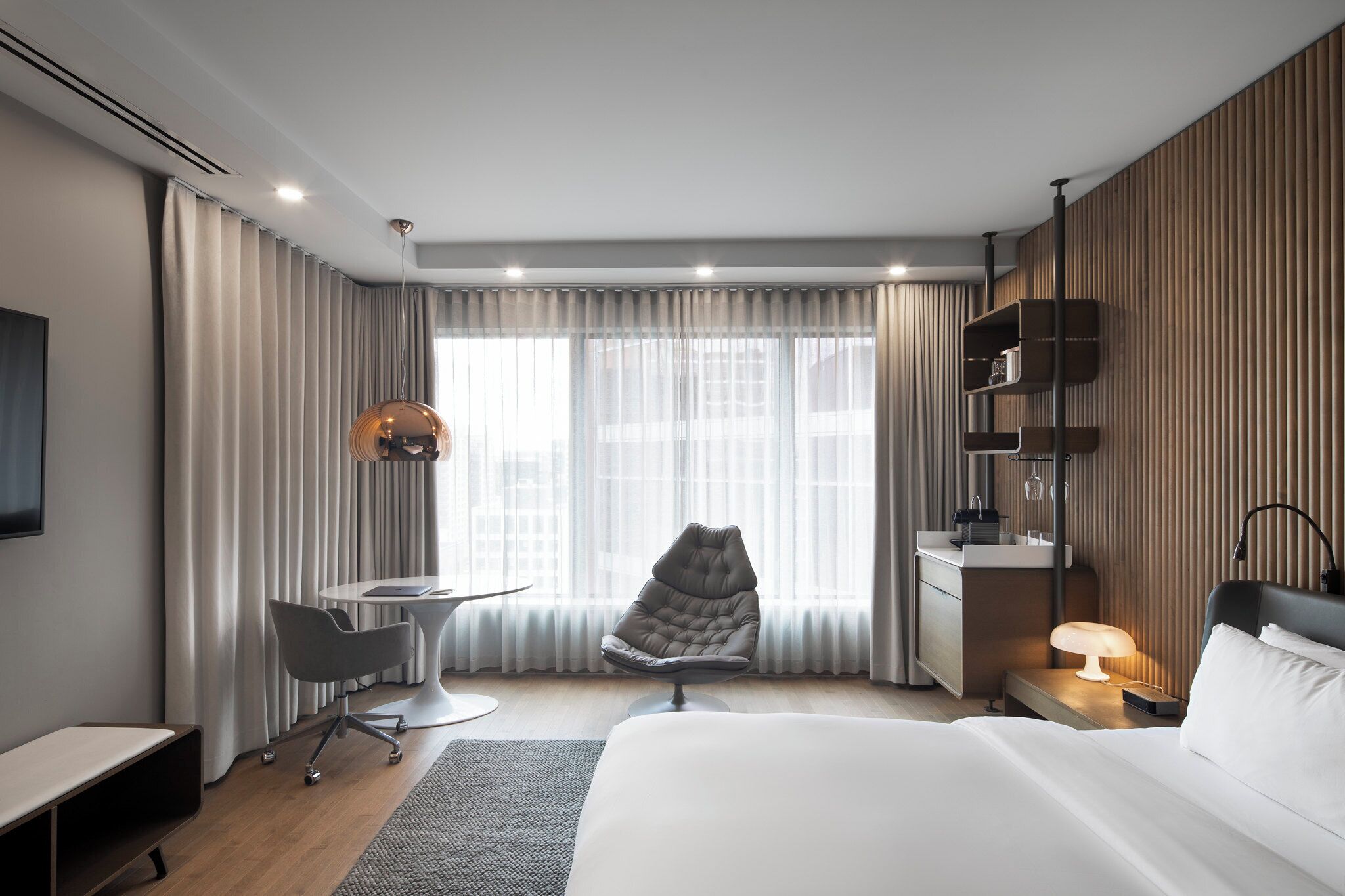 A modern hotel room with a bed, desk, chair, and a view of the city through large windows.