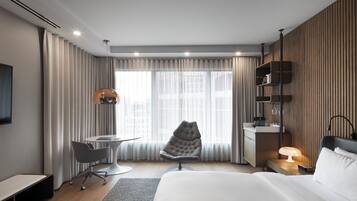 A modern hotel room with a bed, desk, chair, and a view of the city through large windows.