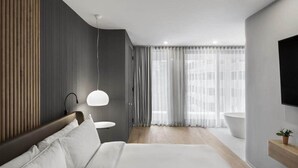 Ein modernes Hotelzimmer mit einem großen Bett, einem Nachttisch, einer Pendelleuchte, einem Badezimmer mit Badewanne und einem großen Fenster mit durchsichtigen Vorhängen.