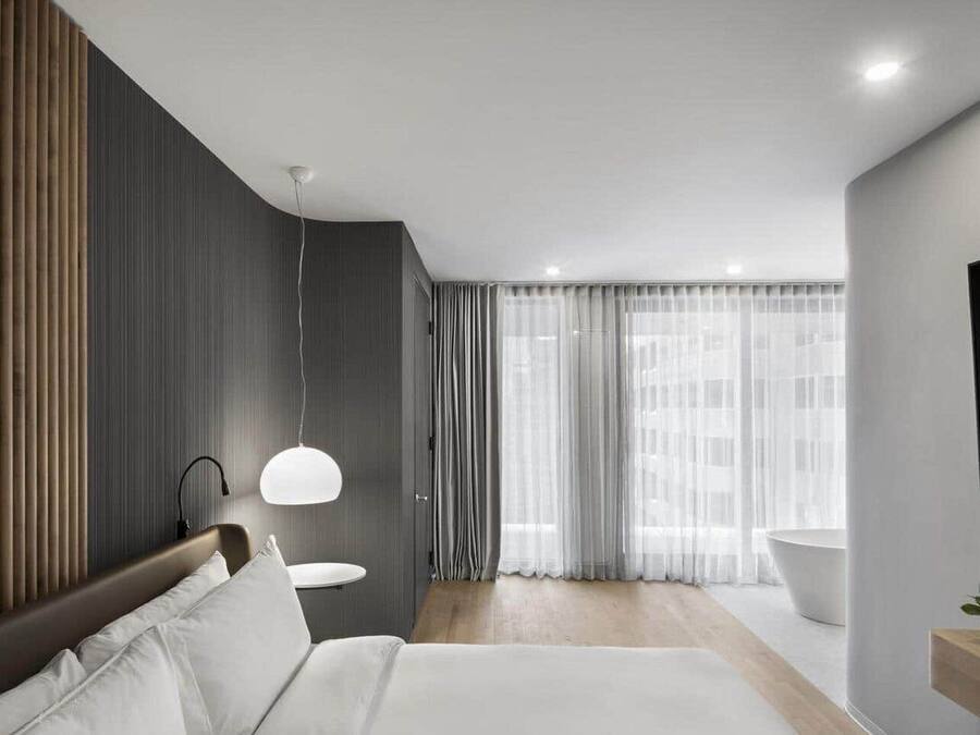 A modern hotel room with a large bed, a bedside table, a hanging light, a bathroom with a bathtub, and a large window with sheer curtains.