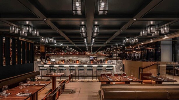 Eine moderne Bar mit verschiedenen Sitzgelegenheiten, darunter Barhocker und Tische, sowie einer zentralen Bar und einer Auswahl an Flaschen und Gläsern.