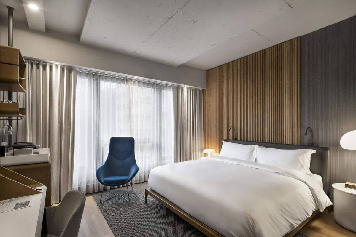 A modern hotel room with a large bed, a desk with a chair, a vanity, and a blue armchair.