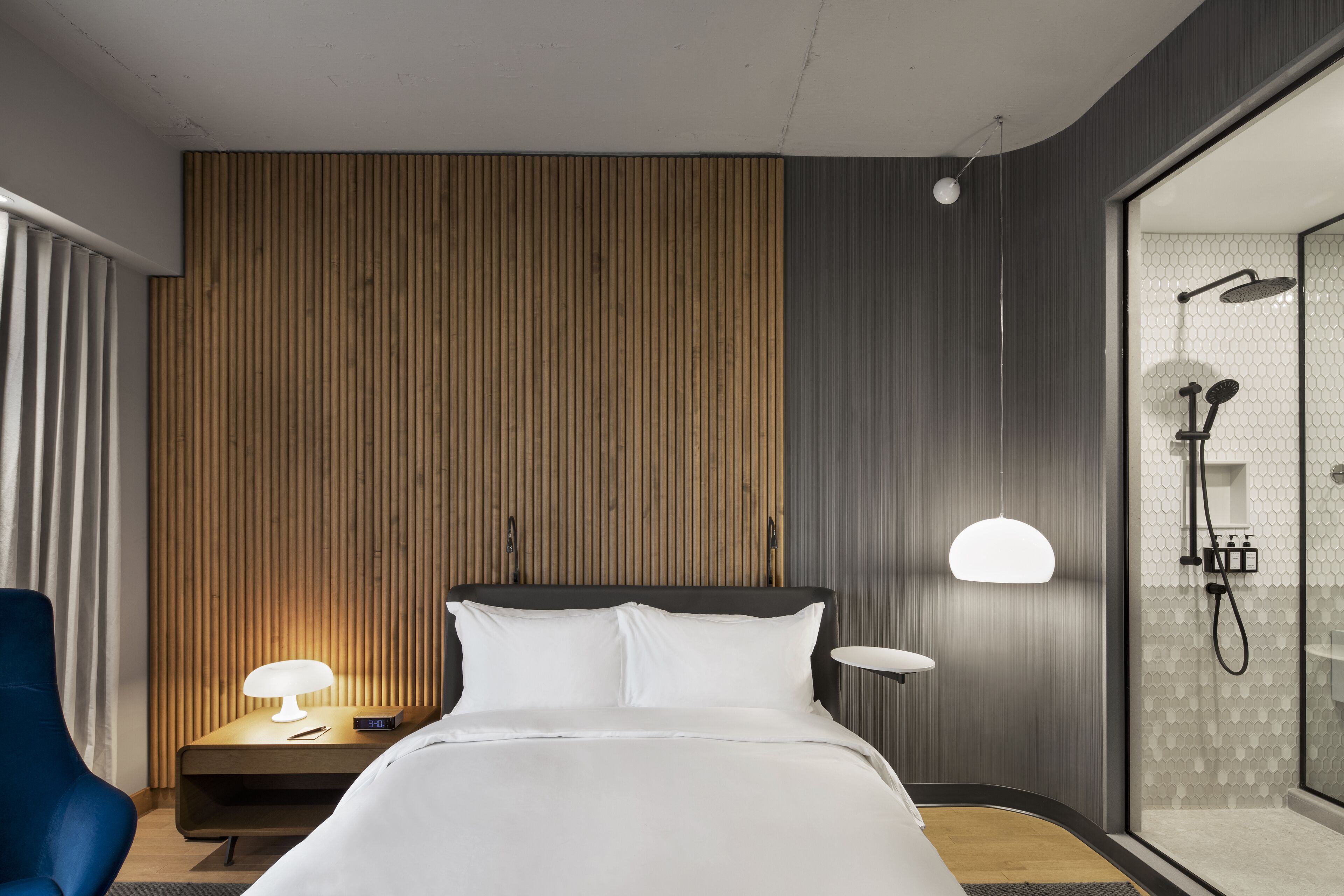Una habitación de hotel moderna con cama, mesita de noche, lámpara y zona de ducha con puerta de cristal.