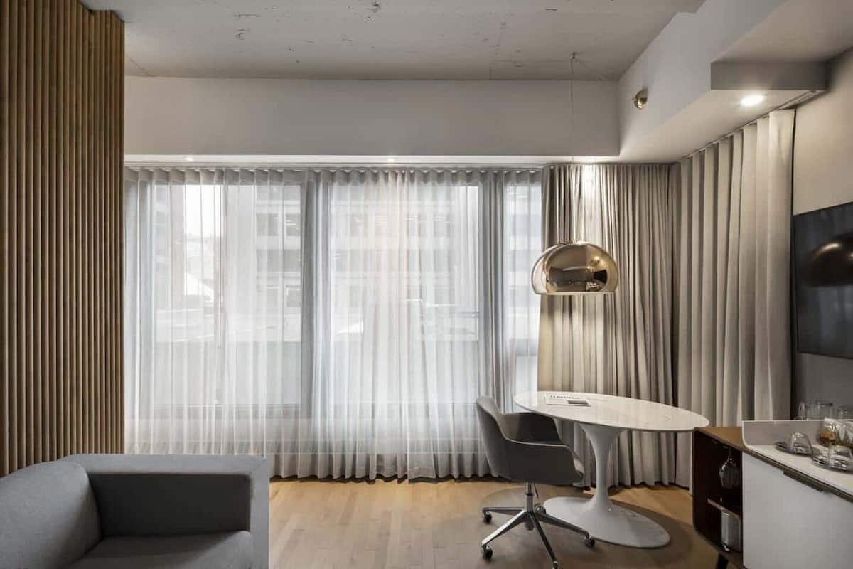 A modern hotel room with a large window, a round table, a sofa, and a TV mounted on the wall.