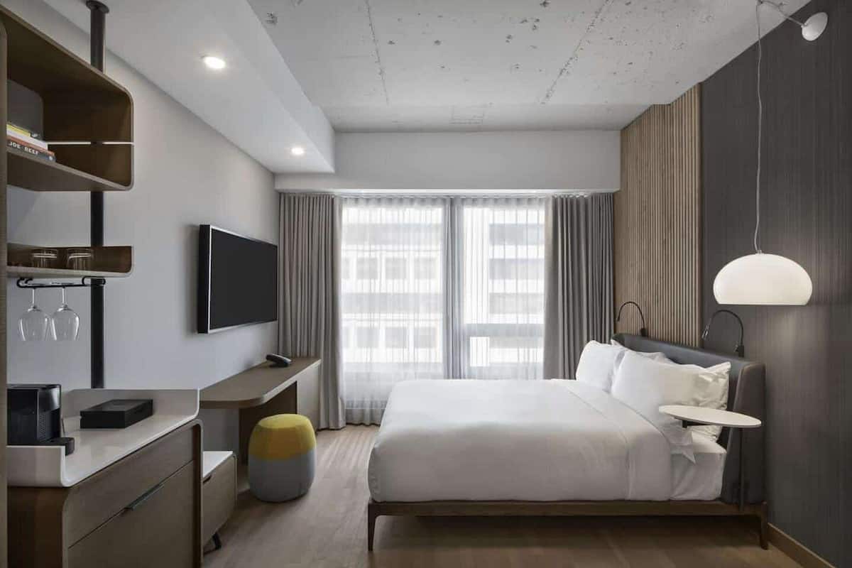 A modern hotel room with a large bed, a desk with a flat-screen TV, a minibar, and a seating area with a lamp.