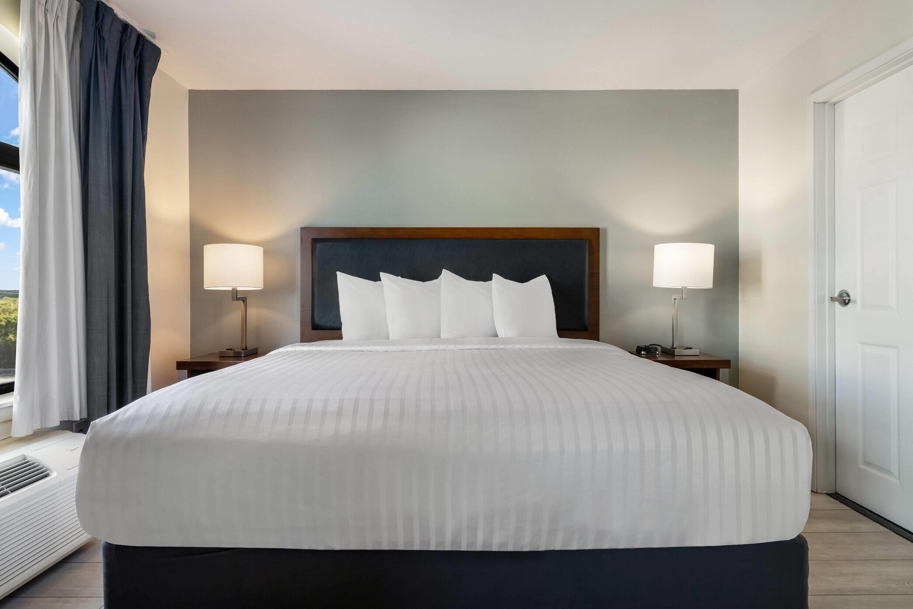 Une chambre d’hôtel avec un grand lit, deux tables de chevet avec des lampes, une vue sur l’extérieur par une fenêtre avec des rideaux, et une porte sur le côté droit.