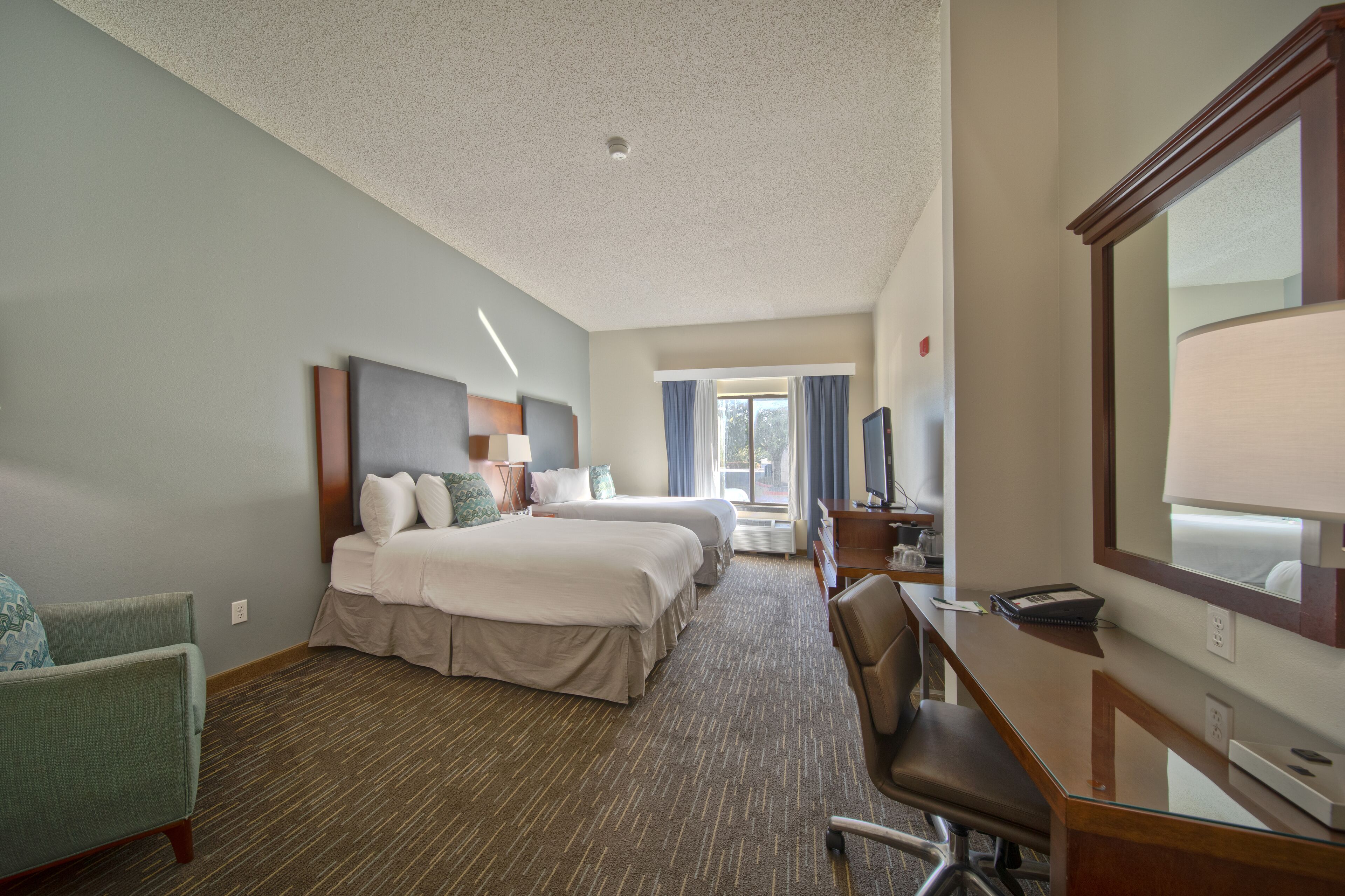 A hotel room with two beds, a desk, a chair, a mirror, and a window with curtains.