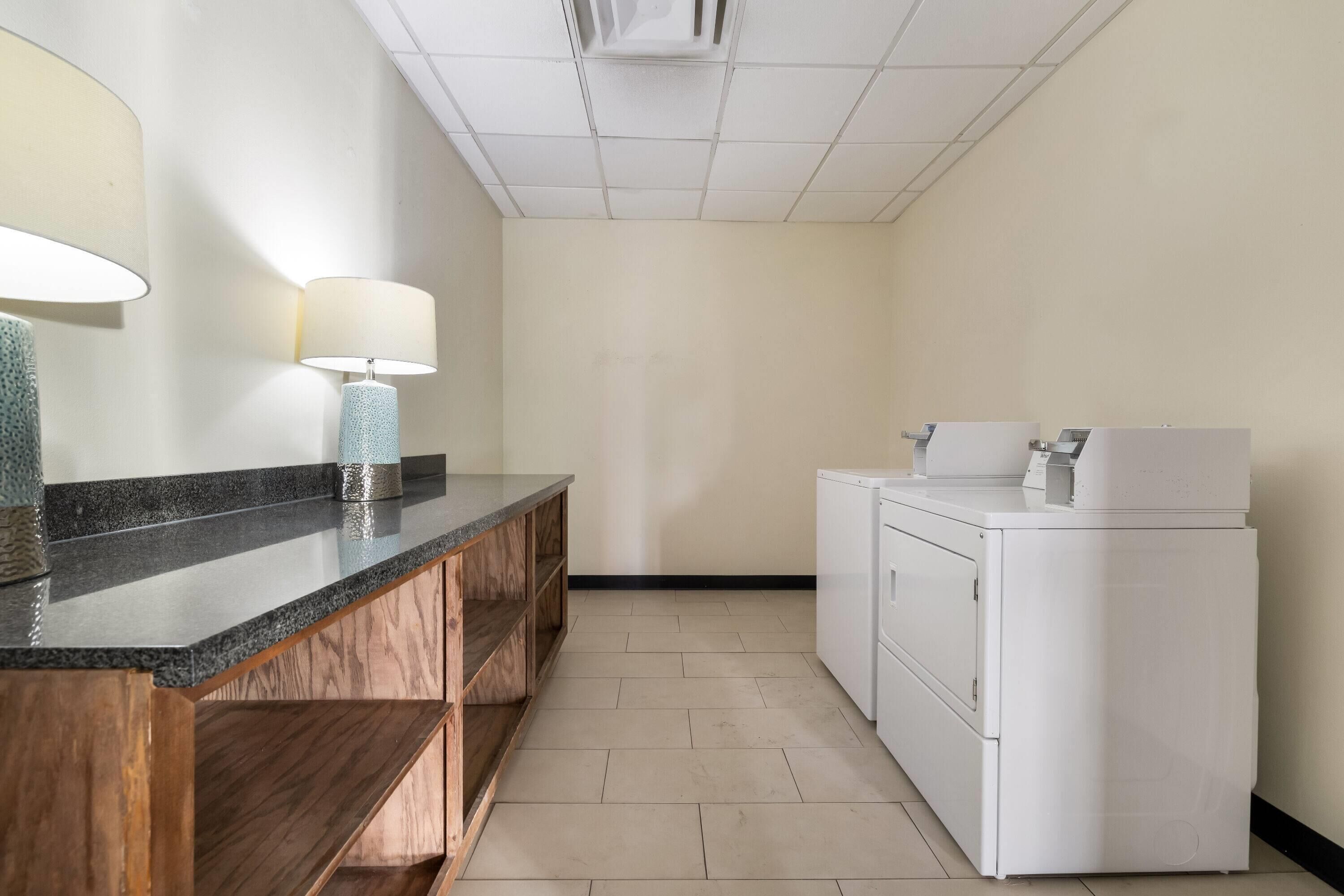 Baño de hotel con encimera de granito, estanterías de madera, lavabo, dos lámparas de pared y lavadora y secadora apiladas.