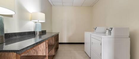 Baño de hotel con encimera de granito, estanterías de madera, lavabo, dos lámparas de pared y lavadora y secadora apiladas.