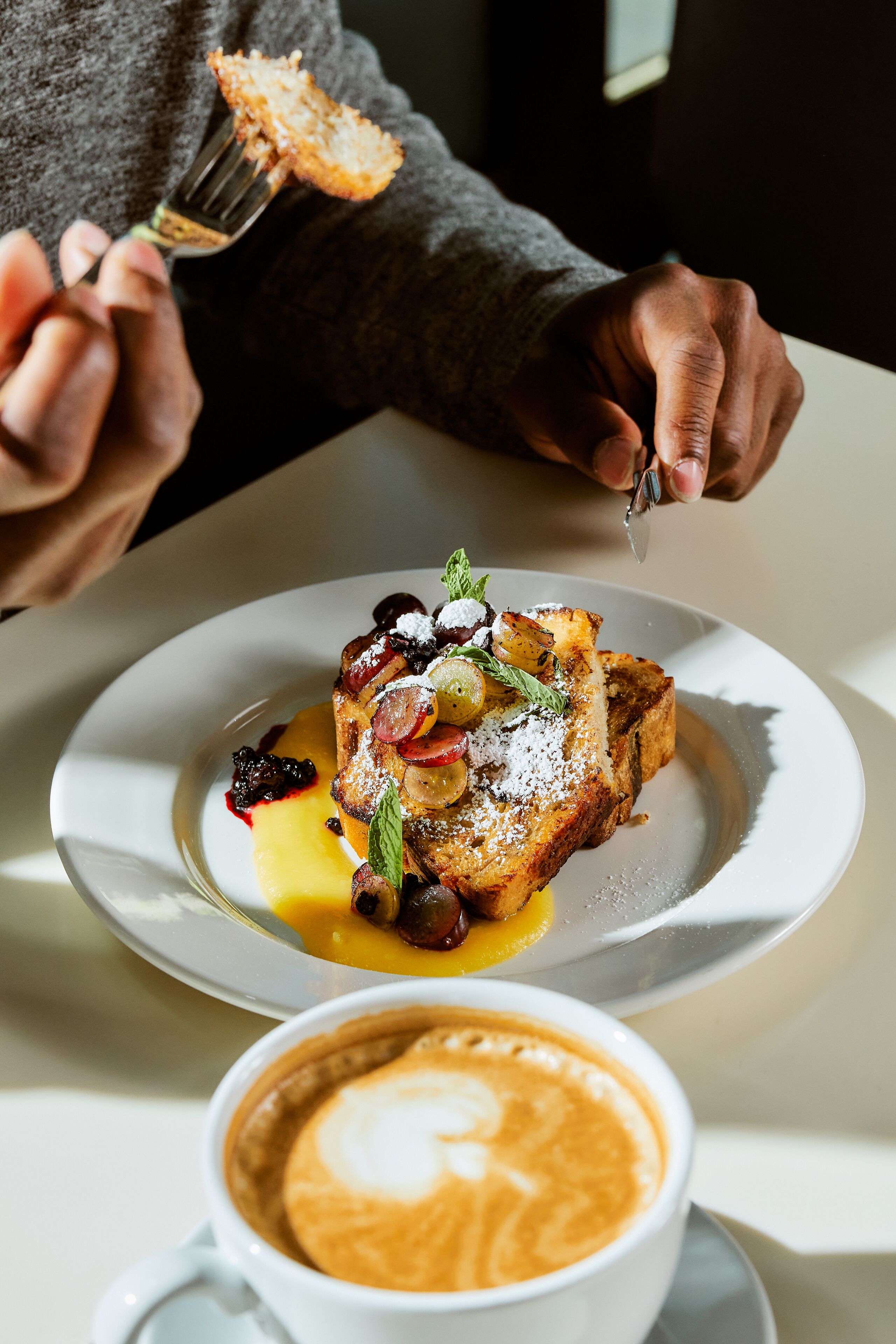 A person holding a fork with a piece of toast topped with fruit and syrup, next to a cup of coffee with latte art.