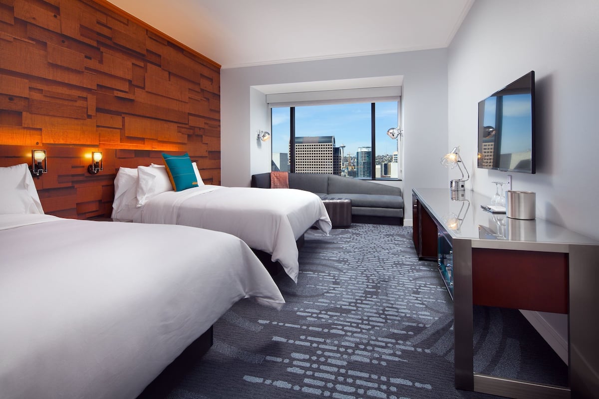 A modern hotel room with two beds, a desk, a sofa, and a large window offering a city view.