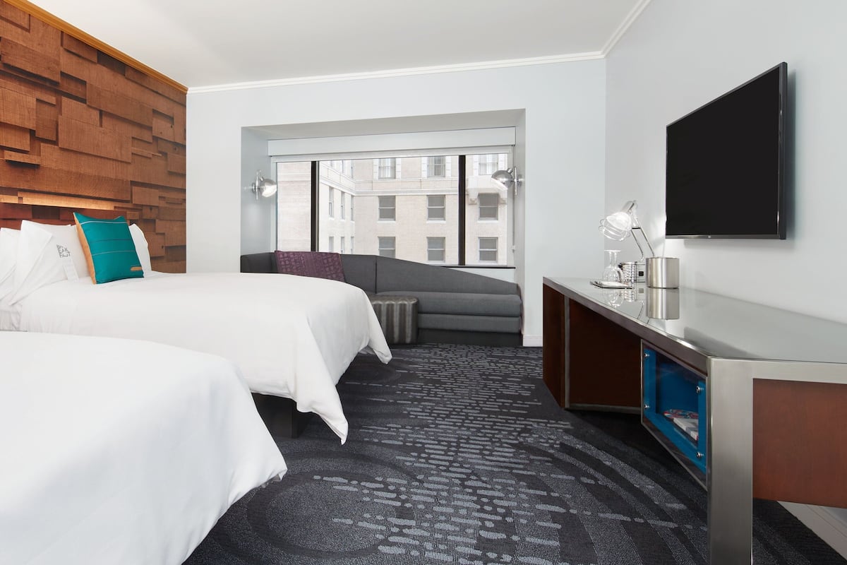 A hotel room with two beds, a desk, a flat-screen TV, and a large window offering a view of buildings.