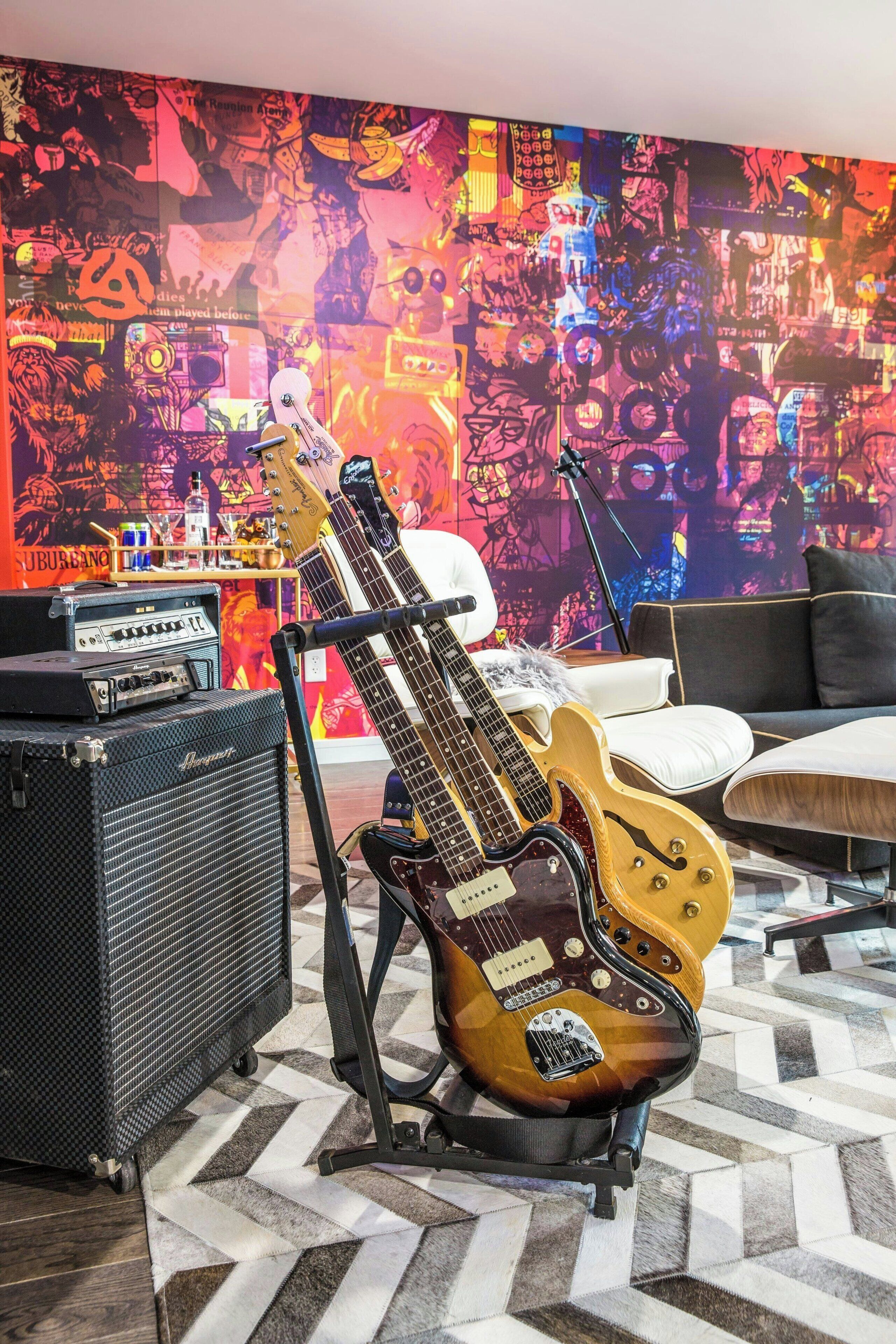 A room with a large, colorful mural on the wall, a black amplifier, a brown electric guitar, and a white couch.