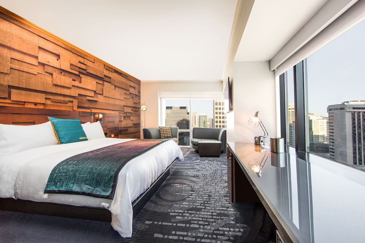 A modern hotel room with a large bed, a city view, a seating area, and a wooden accent wall.