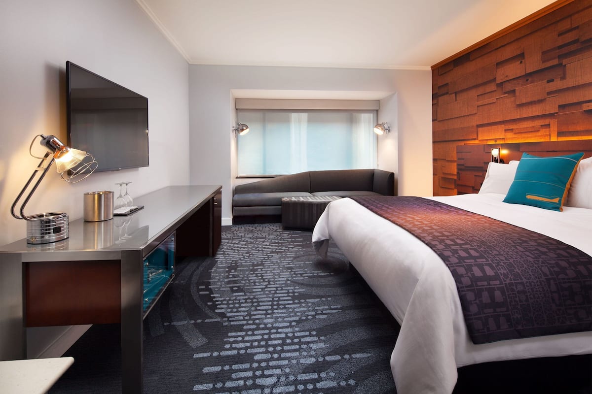 A modern hotel room with a flat-screen TV, a desk with a lamp, a sofa, a bed with a patterned blanket, and a wooden accent wall.