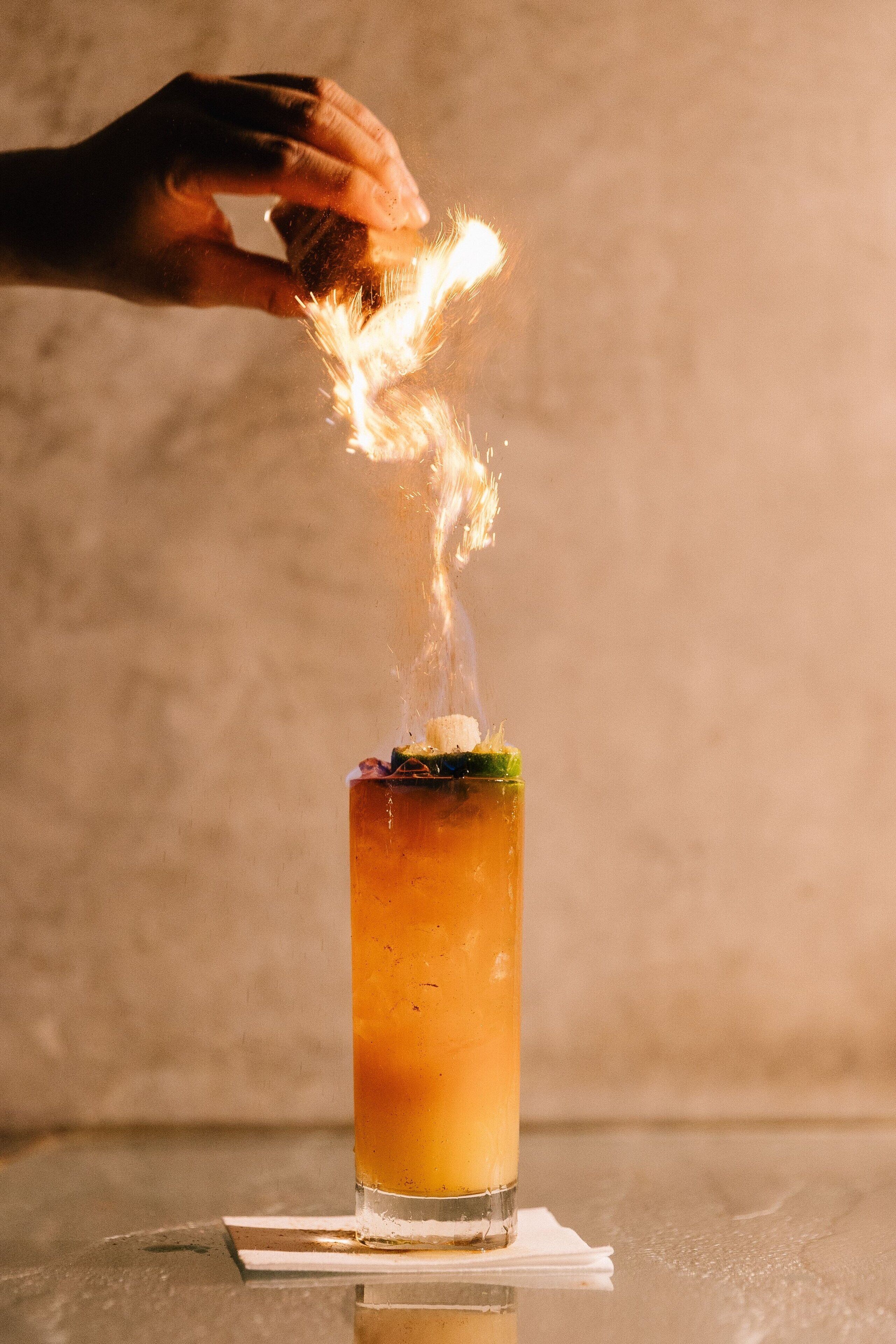 En hand håller en tändsticka ovanför ett högt glas med en vätska i, troligen en cocktail, och allt detta står på en reflekterande yta.