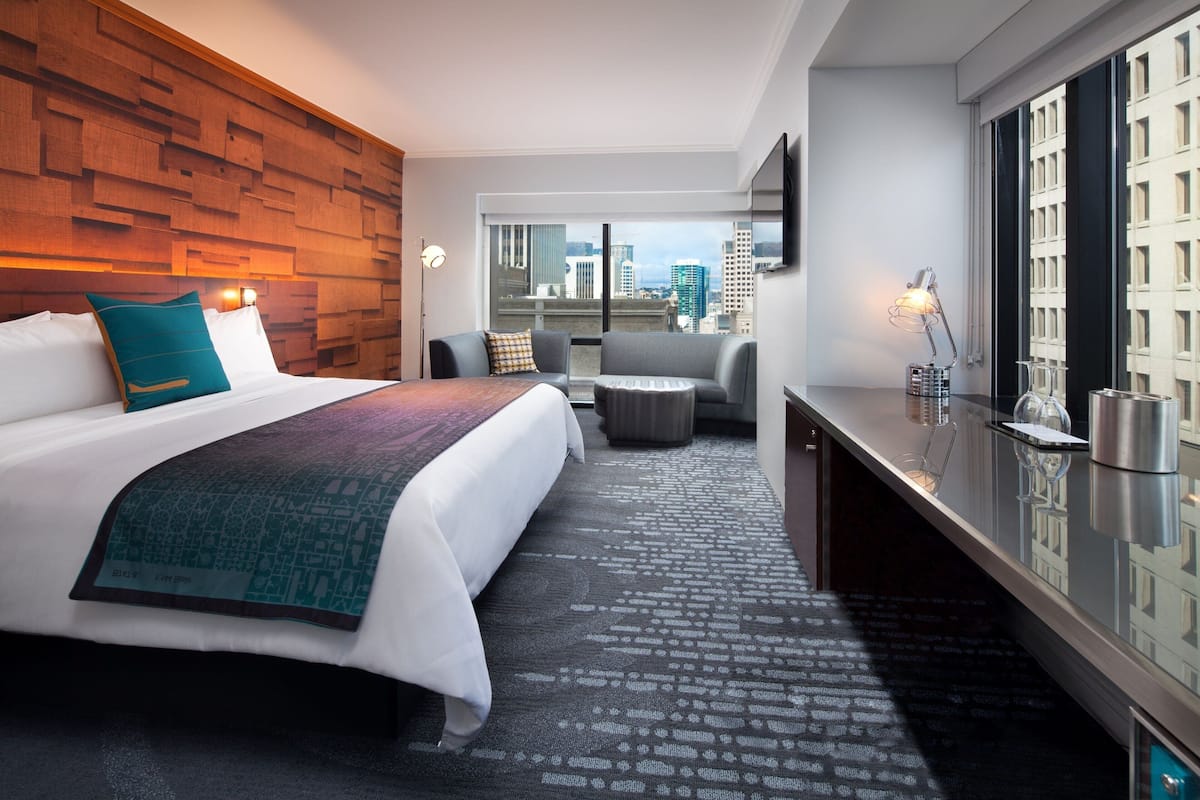 A hotel room with a large bed, a desk with a lamp, a chair, and a view of the city through large windows.