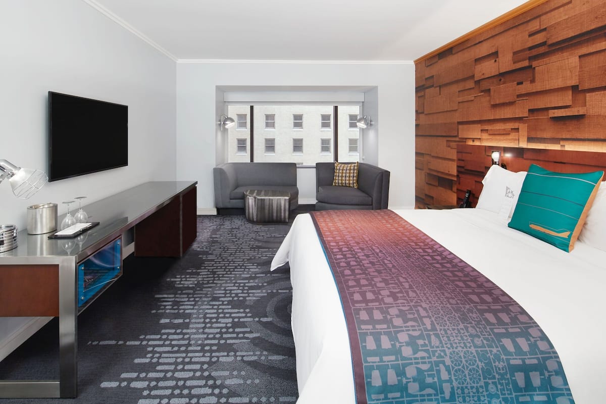 A hotel room with a flat-screen TV, a desk with a lamp, a sofa, a bed with a patterned bedspread, and a wooden headboard.