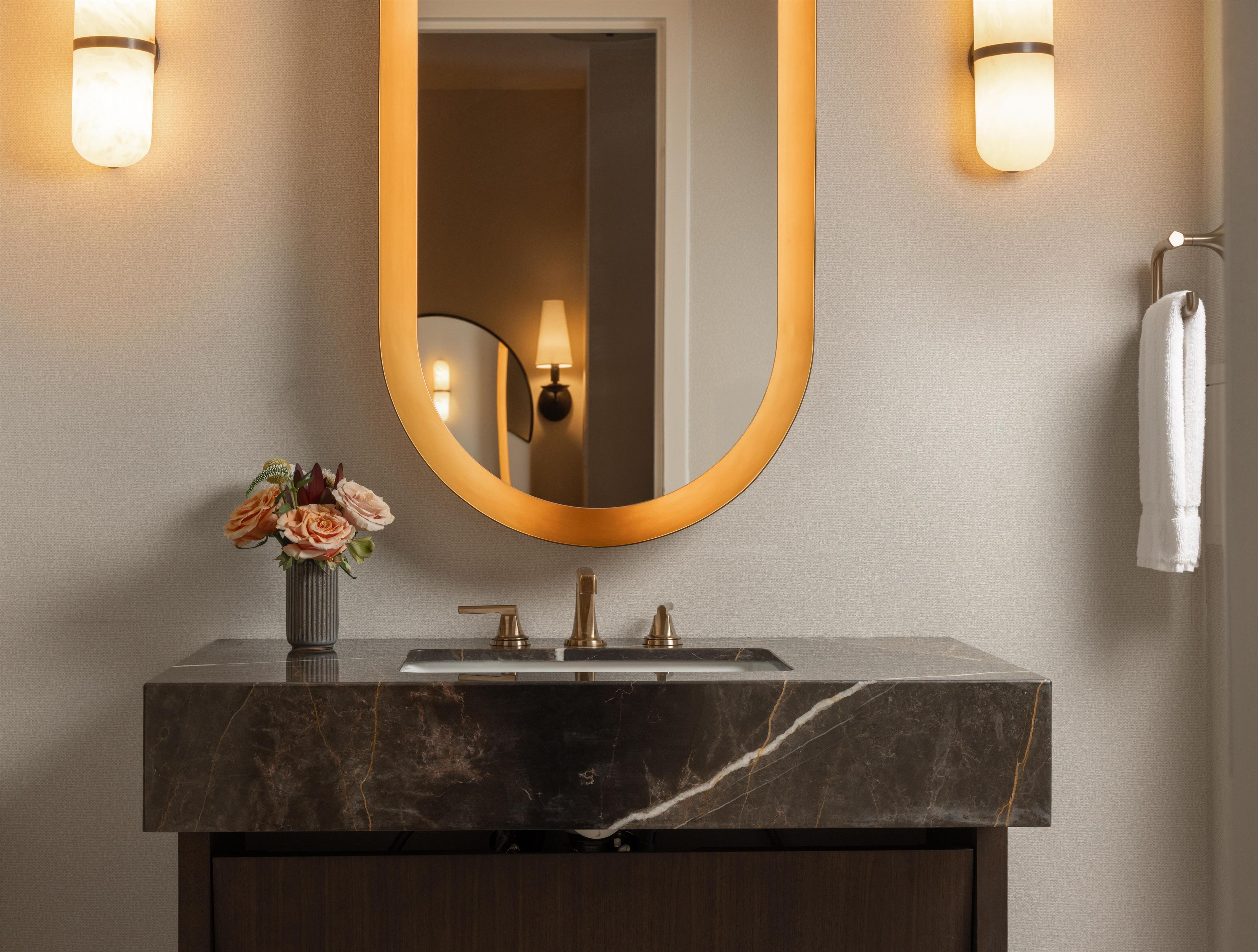 A bathroom with a marble sink, a gold-framed mirror, and a vase of flowers.